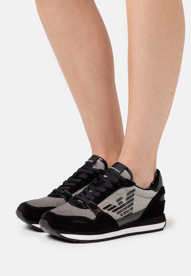 Jalka, jotka käyttävät mustia ja hopeita matalalattiaisia tennareita logo-suunnittelulla, seisovat valkoista taustaa vasten.