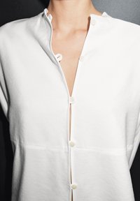 Hvid skjorte med knapper, der har en høj krave, vandrette syninger og en knapliste med lyse knapper, lavet af et blødt, glat stof.