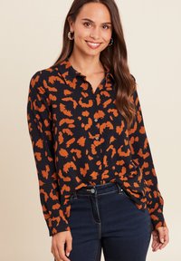 Frau, die lächelt und ein schwarzes Hemd mit orangefarbenem Leopardenmuster und dunkelblauen Jeans trägt, vor einem beigen Hintergrund.