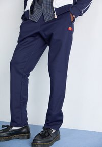 Marineblauwe sweatpants van een gladde stof, met een klein rood en blauw logo, gecombineerd met zwarte schoenen met een kwastdetail en dikke zolen.