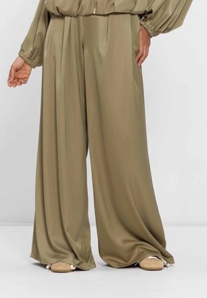 Persoon draagt losse, wijde, olijfgroene satijnen broek en beige instappers, staand tegen een effen lichte achtergrond.