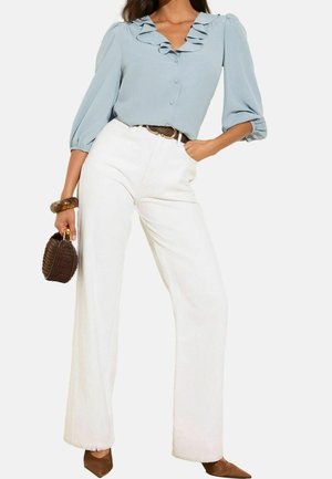 Femme portant une blouse bleu clair à volants, un pantalon blanc taille haute large, une ceinture marron, tenant un sac à main tressé marron foncé.