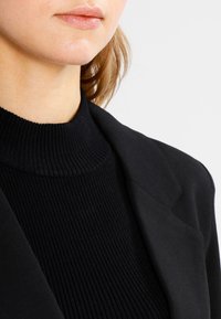 Svart ribbad polotröja i lager under en skräddarsydd svart blazer, med strukturerade axlar och en mjuk tygkvalitet.