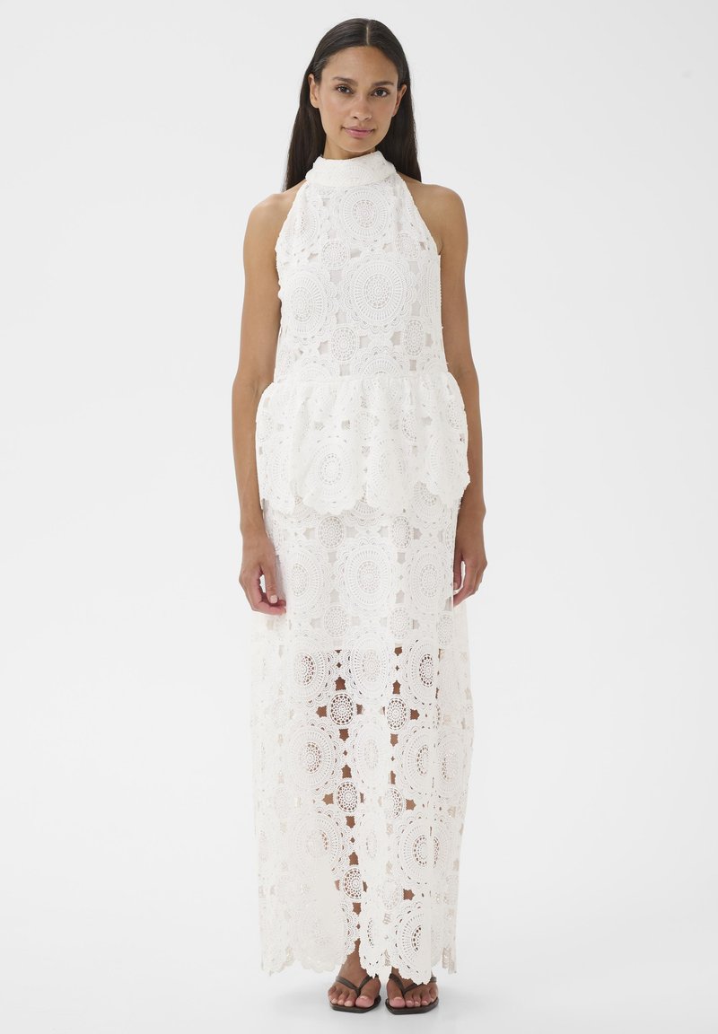 Femme portant une robe blanche en dentelle crochetée sans manches avec un col montant et un ourlet festonné, debout devant un fond uni.