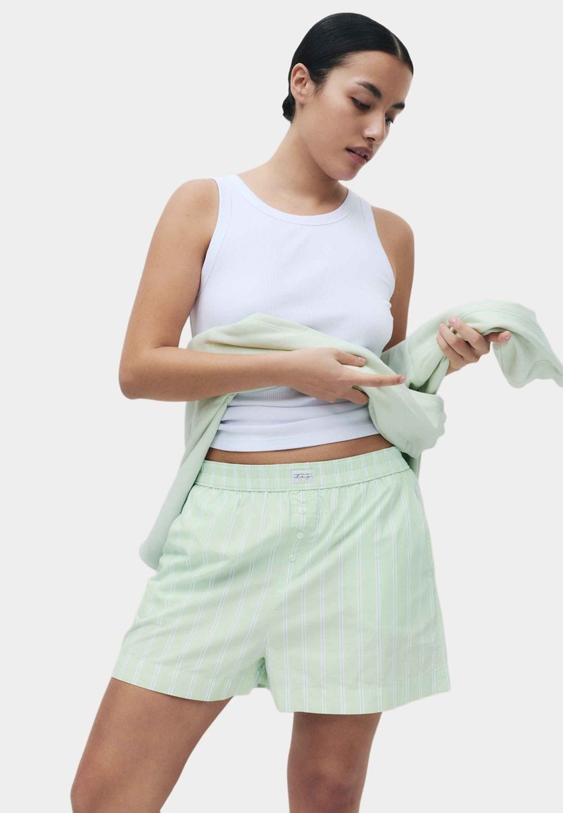 Hellgrüne gestreifte Shorts aus weichem Stoff mit lockerem Schnitt. Kombiniert mit einem weißen Tanktop und hält ein hellgrünes Kleidungsstück in der Hand.