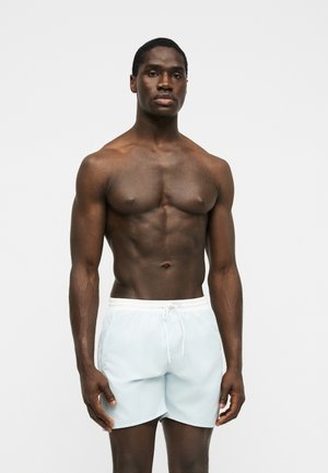Uomo muscoloso in piedi, indossa pantaloncini da bagno azzurri con coulisse bianca su uno sfondo chiaro e uniforme.