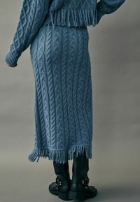 Fustă lungă tricotată, de culoare albastră, cu margine franjurată, asociată cu bocanci negri cu talpă groasă, având accente de cataramă. Materialul texturat evidențiază modele complexe.