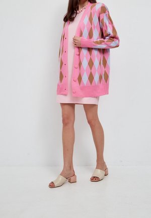 Femme portant un cardigan à motifs losanges rose et marron sur une robe rose clair, chaussures à talons blocs ouvertes beige, posant devant un fond blanc.
