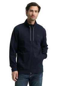 Marineblauwe jas met ritssluiting van een gestructureerde stof, voorzien van een hoge kraag, verstelbare trekkoorden en twee zijzakken, gedragen met denim jeans.