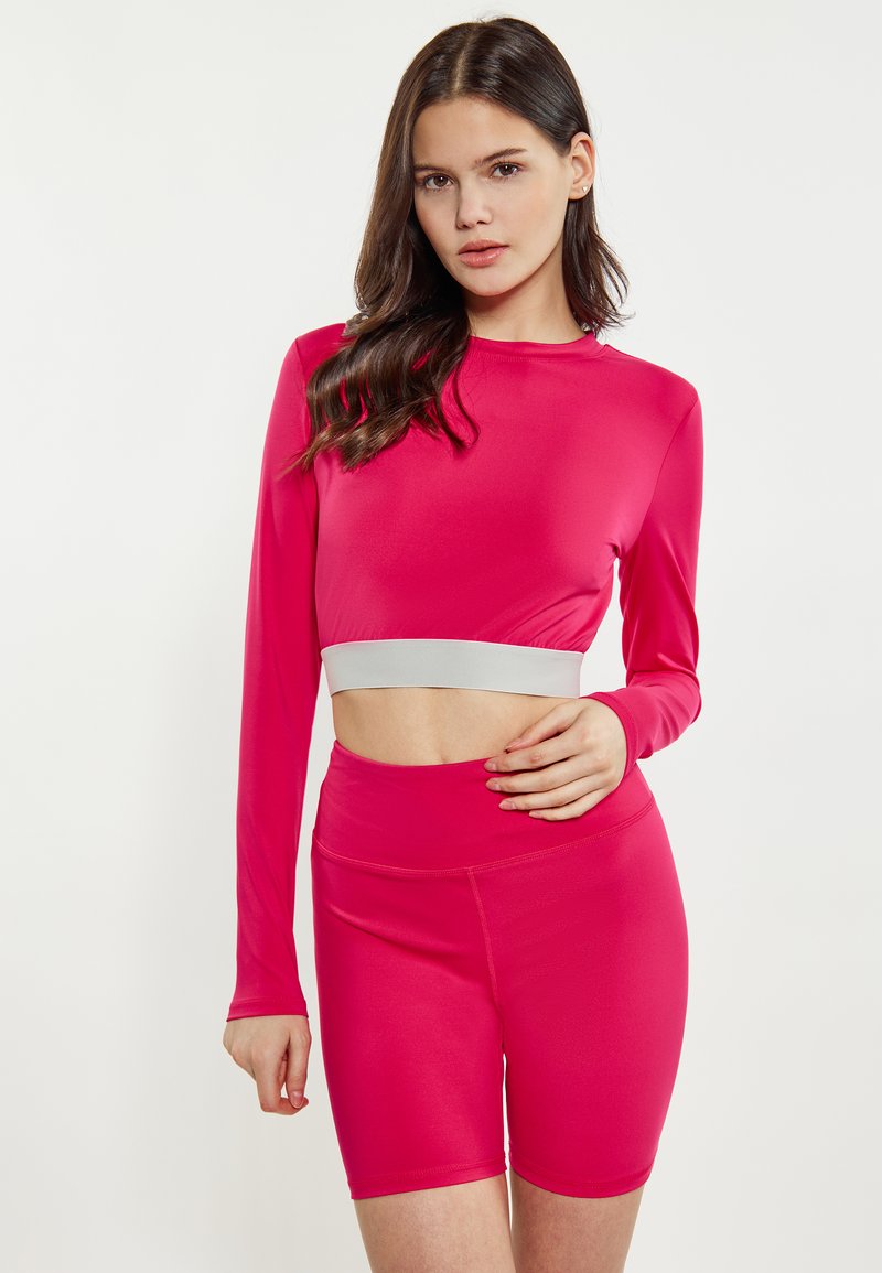 Set de îmbrăcăminte activă ajustat în roz vibrant. Top scurt cu mâneci lungi și un band elastic gri, împreună cu pantaloni scurți cu talie înaltă. Material neted și elastic.