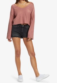 Top corto de manga larga de color rosa empolvado texturizado combinado con pantalones cortos de mezclilla negros de cintura alta. La modelo lleva zapatillas blancas.