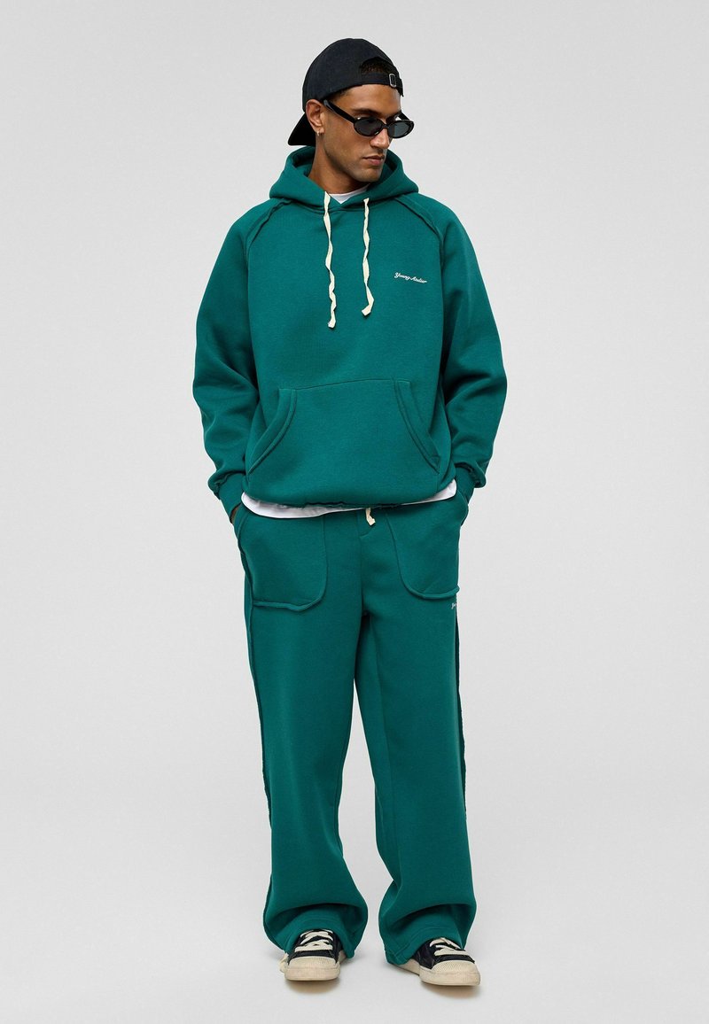Teal fleece hoodie met trekkoord, grote frontzak en bijpassende wijde broek. Gedragen met een zwarte pet en zonnebril.