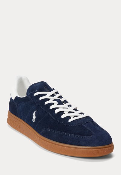 Tamsiai mėlynas zomšinis sportinis batelis su baltais raišteliais, baltu kulno apvadu, rudu guminiu padu ir baltu Polo Ralph Lauren logotipu šone.