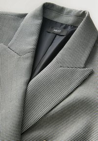 Gros plan sur un blazer à motif pied-de-poule noir et blanc avec une doublure intérieure en satin noir et une étiquette portant l’inscription NEXT.