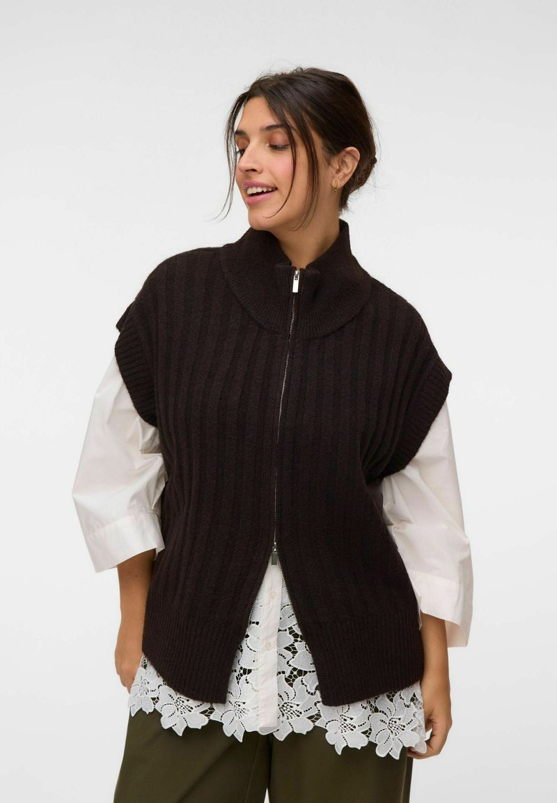 Femme portant un pull sans manches noir côtelé avec fermeture éclair sur une chemise blanche bordée de dentelle et un pantalon vert olive, regardant légèrement sur le côté.