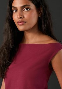 Bordo blūze bez pleciem ar īsām piedurknēm. Gluda, elastīga auduma, pieguļošs dizains ar diskrētu, tīru kakla izgriezumu.