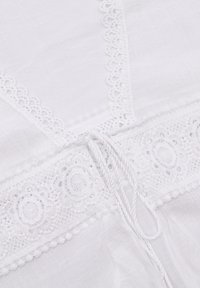 Indumento bianco con delicata rifinitura in pizzo, tessuto texturizzato, dettaglio con cordoncino annodato e accenti di uncinetto floreale in vita.