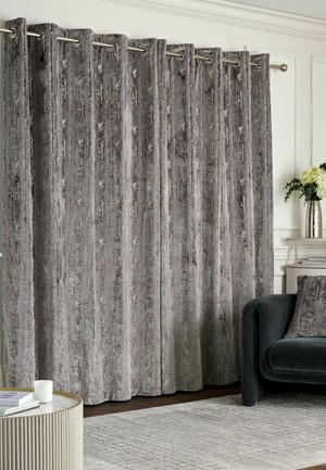 Gray textured curtains hanging from a silver rod in a living room with a dark sofa, patterned rug, round table, and vase with flowers.