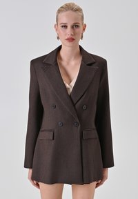 Blazer marrone doppiopetto con ampi rever, due tasche frontali e tessuto testurizzato. Presenta bottoni neri e un taglio più corto.