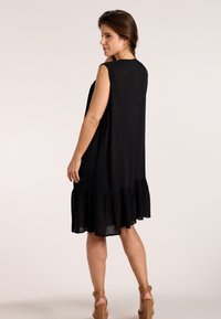 Ženska oblečena v črno obleko brez rokavov do kolen z nabrano spodnjo robno plastjo, stoji bočno, obuta v rjave tkanine platformne sandale.