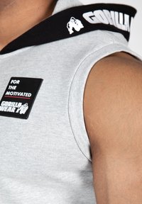 Grijze mouwloze hoodie met een zwarte capuchon, voorzien van een rubberen patch met de tekst "VOOR DE GEMOTIVEERDEN" en het "GORILLA WEAR" logo in het wit.