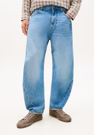 Osoba ubrana w jasnoniebieskie luźne dżinsy, brązowe trampki i brązową kratowaną koszulę, z jedną ręką w kieszeni.