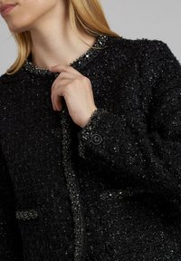 Schwarze strukturierte Jacke mit Perlenakzenten, rundem Ausschnitt und Knopfverschlüssen. Sie verfügt über einen weichen Stoff mit einem leicht strukturierten Schnitt.
