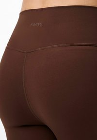 Smeđe tajice napravljene od glatkog, rastezljivog materijala. Imaju visoki pojas s imenom branda "PIECES" utisnutim blizu vrha.