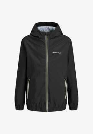 Junge mit weißer Mütze, schwarzer Zip-up-Jacke, hellgrünem T-Shirt, lockeren schwarzen Jeans und weißen Turnschuhen, der steht und nach links schaut.