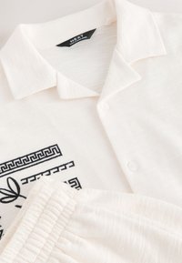 Camisa blanca con cuello, estampado geométrico y de hojas negras, y pantalones blancos con cintura elástica a juego.
