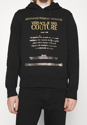 Personne portant un sweat à capuche noir Versace Jeans Couture avec un texte doré et un motif de chaîne, pantalon noir, debout les bras détendus.