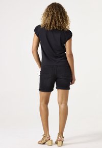 Czarny top z krótkim rękawem w połączeniu z ciemnymi szortami jeansowymi z podwiniętymi brzegami i tylnymi kieszeniami. Modelka ma na sobie złote sandały na słupku.