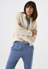 Frau in einer cremefarbenen, flauschigen Jacke, einem bunten Pullover und blauen Cordhosen, die die Arme verschränkt hält und leicht zur Seite blickt, vor einem weißen Hintergrund.