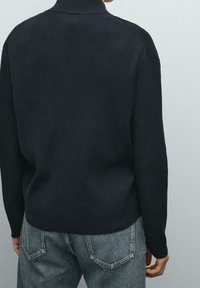 Henkilö, jolla on päällään musta ribbipaita olkataskuilla ja haalistuneet siniset farkut, näkyy selkäpuolelta vaaleaa taustaa vasten.