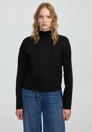 Black, long-sleeve turtleneck top with a relaxed fit and minimal seams, paired with light blue wide-leg jeans. Smooth fabric texture.