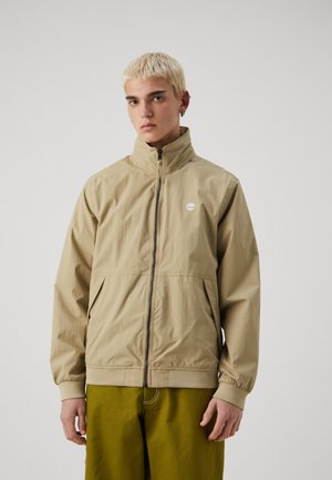 Giacca beige con zip e colletto alto, polsini elastici e un emblema con logo. Tessuto liscio e materiale leggero, abbinata a pantaloni verde oliva.