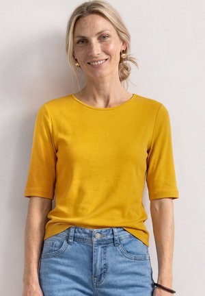 Femme blonde souriante portant des boucles d'oreilles créoles dorées, un top manches courtes jaune moutarde, et un jean denim bleu clair sur un fond uni.