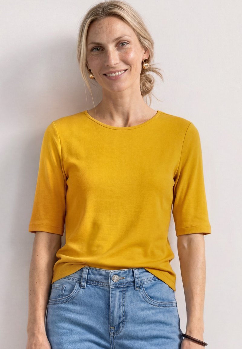 Femme blonde souriante portant des boucles d'oreilles créoles dorées, un top manches courtes jaune moutarde, et un jean denim bleu clair sur un fond uni.