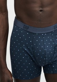 Αρσενικός κορμός που φοράει μπλε σκούρα μποξεράκια με ανοιχτό μπλε διαμαντένιο μοτίβο και λογότυπο GAP στη μέση, σε λευκό φόντο.