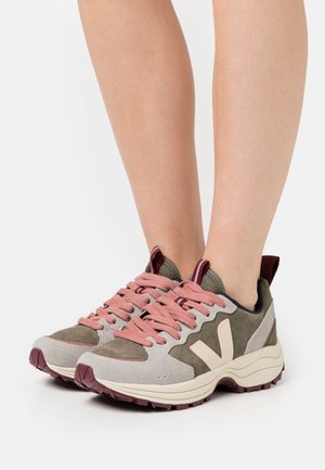 Tenis cinza claro e verde oliva com atacadores rosa, materiais em camurça e têxtil, solas em borracha grossa e um logótipo em destaque de lado.