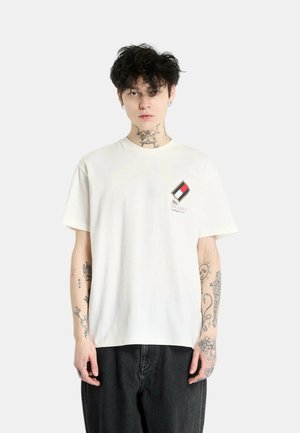 Camiseta blanca de algodón con un pequeño parche multicolor en el lado izquierdo del pecho. Modelo vistiendo jeans oscuros, con tatuajes visibles en los brazos.