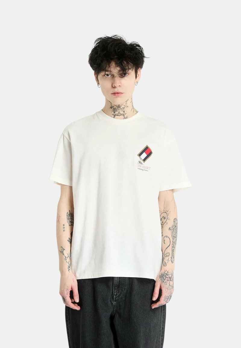 Camiseta blanca de algodón con un pequeño parche multicolor en el lado izquierdo del pecho. Modelo vistiendo jeans oscuros, con tatuajes visibles en los brazos.