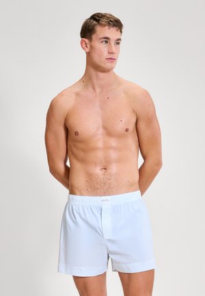 Uomo senza maglietta con capelli castano corti che indossa boxer a righe azzurro chiaro, in piedi con le mani dietro la schiena, che guarda di lato.