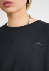 Juodas medvilninis džemperis su apvalia kaklo iškirpte, su iškilmiu Adidas logotipu kairėje krūtinės pusėje. Minkšta medžiaga ir laisvas siluetas.