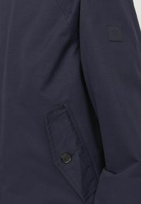 Chaqueta de tela azul marino con un bolsillo frontal con botón, textura suave y un parche de logo cuadrado en la manga.