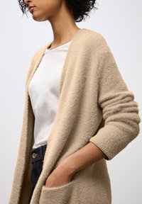 Cardigan lungo beige e testurizzato con apertura frontale e maniche arrotolate, indossato sopra una camicia bianca, con le mani nelle tasche di jeans scuri.