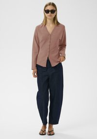 Ein rostfarbener Cardigan mit V-Ausschnitt, Knopfleiste und langen Ärmeln, kombiniert mit locker sitzenden dunkelblauen Jeans und schwarzen Sandalen.