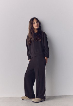 Jeune femme aux longs cheveux portant un survêtement noir ample avec des bandes blanches latérales et des chaussures beiges sans lacets, debout contre un mur gris clair.