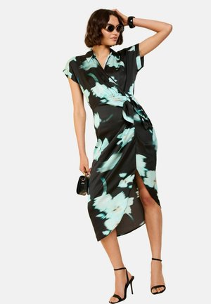 Zwart bloemen wikkeljurk met turquoise patronen, korte mouwen, een zijsplit en een geknoopte detail in de taille. Combinatie met zwarte gespenhakken.