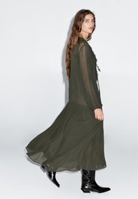 Langes, olivgrünes Kleid aus leichtem Stoff mit Rüschendetails vorne und langen Ärmeln, kombiniert mit schwarzen Stiefeletten.